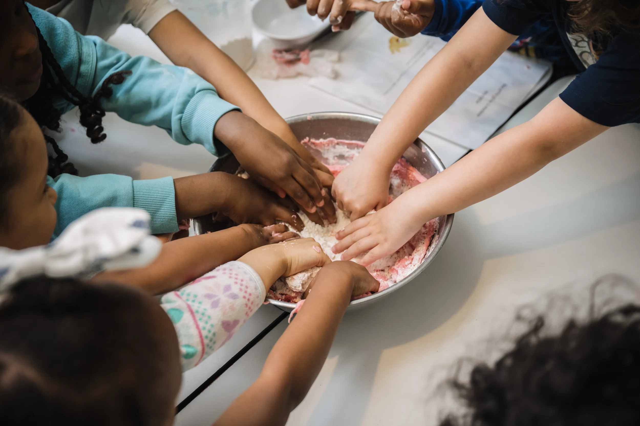 Plusieurs enfants plongent leurs mains dans un grand saladier en métal rempli d'un mélange de farine et de pâte rose, s'adonnant ensemble à une activité amusante et salissante autour d'une table blanche.