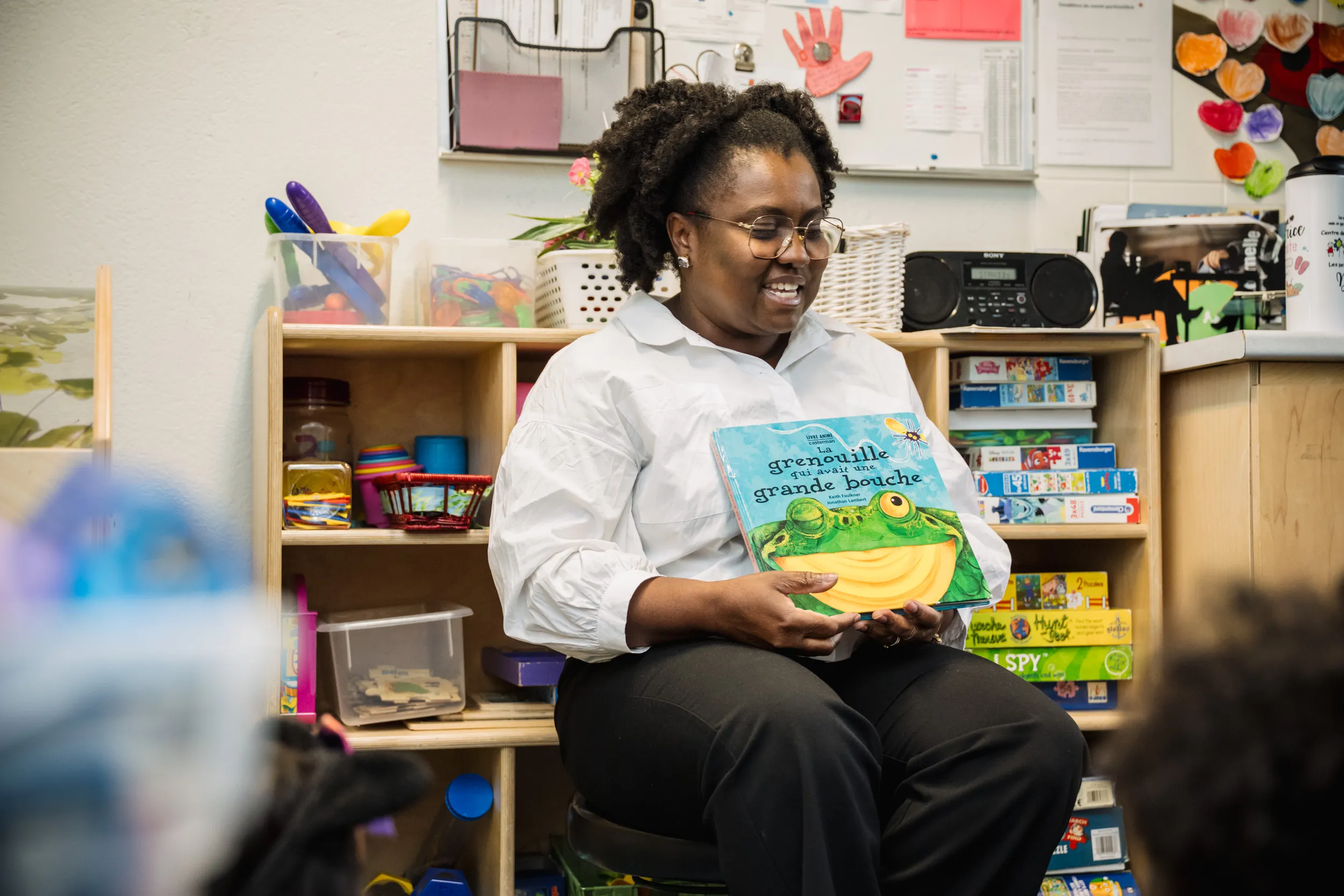 Une femme portant des lunettes et une chemise blanche est assise dans une salle de classe ; elle sourit en tenant un livre pour enfants intitulé *Grenouille à grande bouche* et en faisant la lecture à un groupe d'enfants.