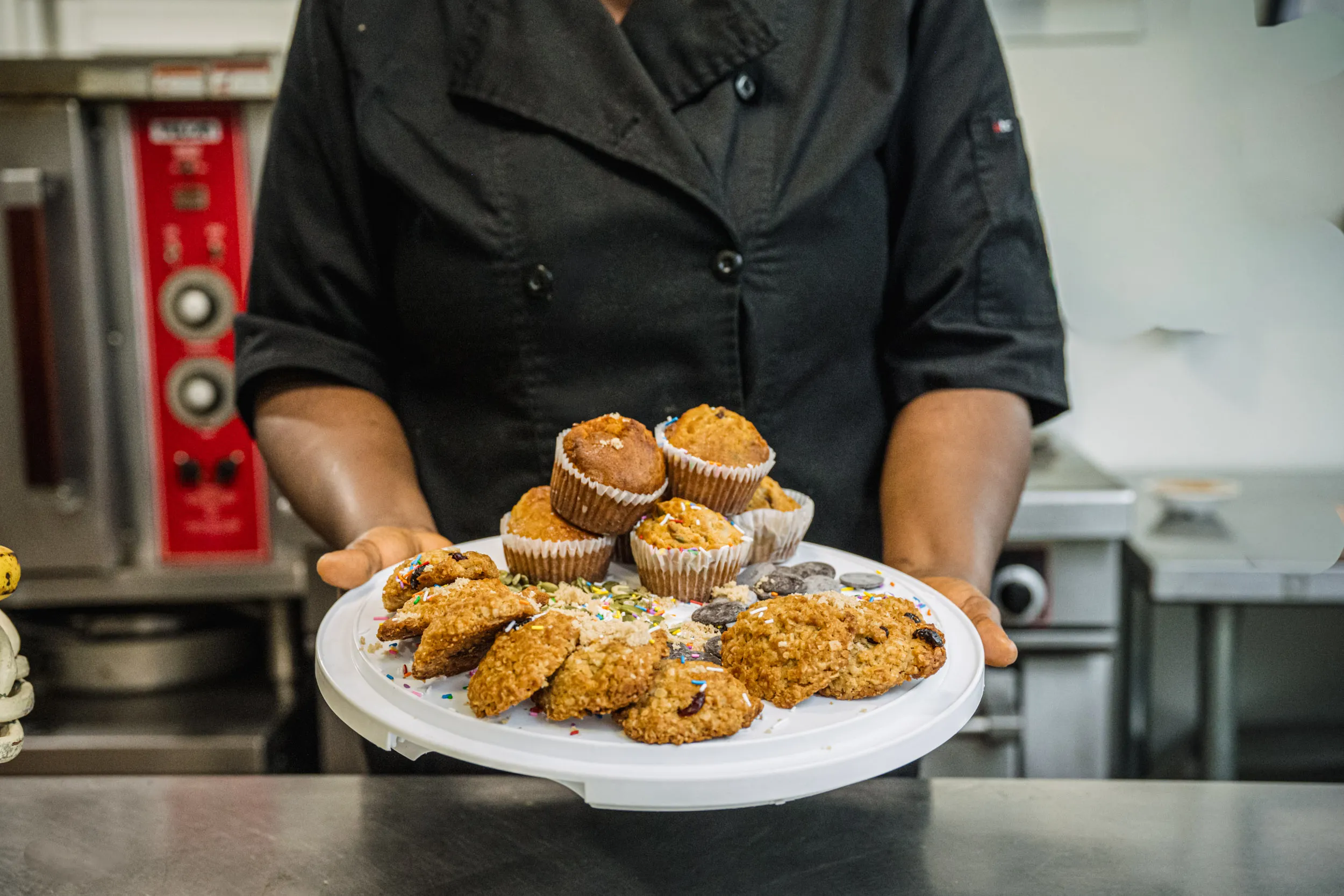 Dans une cuisine professionnelle, une personne vêtue d'un uniforme de chef noir tient un plateau blanc sur lequel sont disposés divers produits de pâtisserie, notamment des biscuits et des muffins.