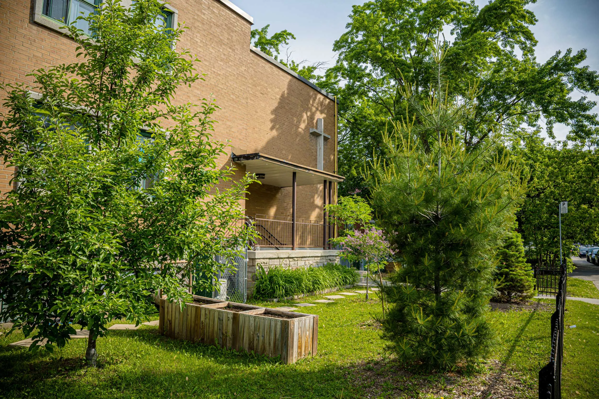Une maison en briques, dotée d'un auvent métallique, est entourée d'arbres verdoyants, de pelouse et d'une jardinière en bois dans la cour. Une clôture noire est partiellement visible sur le côté droit.