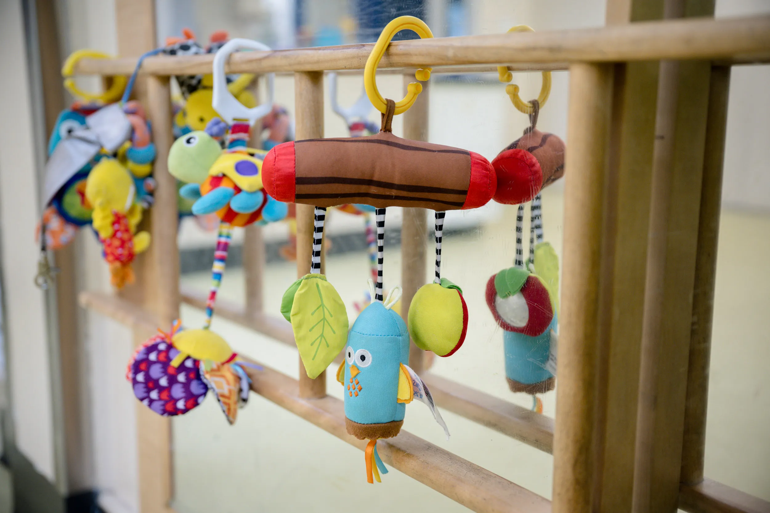 Des jouets souples et colorés pour bébés sont suspendus à la barre en bois d'un lit de bébé : on y trouve notamment un oiseau en peluche, des feuilles et d'autres formes d'animaux ornées de miroirs et de motifs, qui se reflètent dans la vitre située derrière.