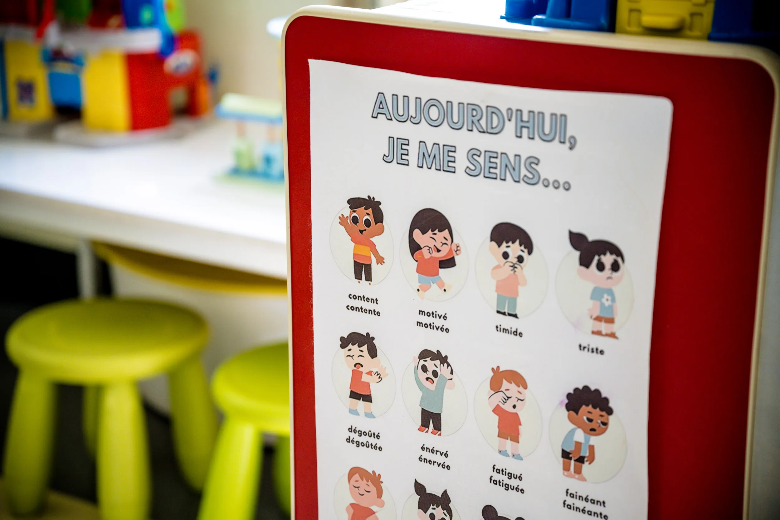 Une affiche en français représentant des enfants sous forme de personnages de bande dessinée exprimant différentes émotions, telles que la joie, la motivation, la timidité et la tristesse, est accrochée dans une salle de classe où l'on aperçoit, en arrière-plan, des tabourets verts et des jouets.