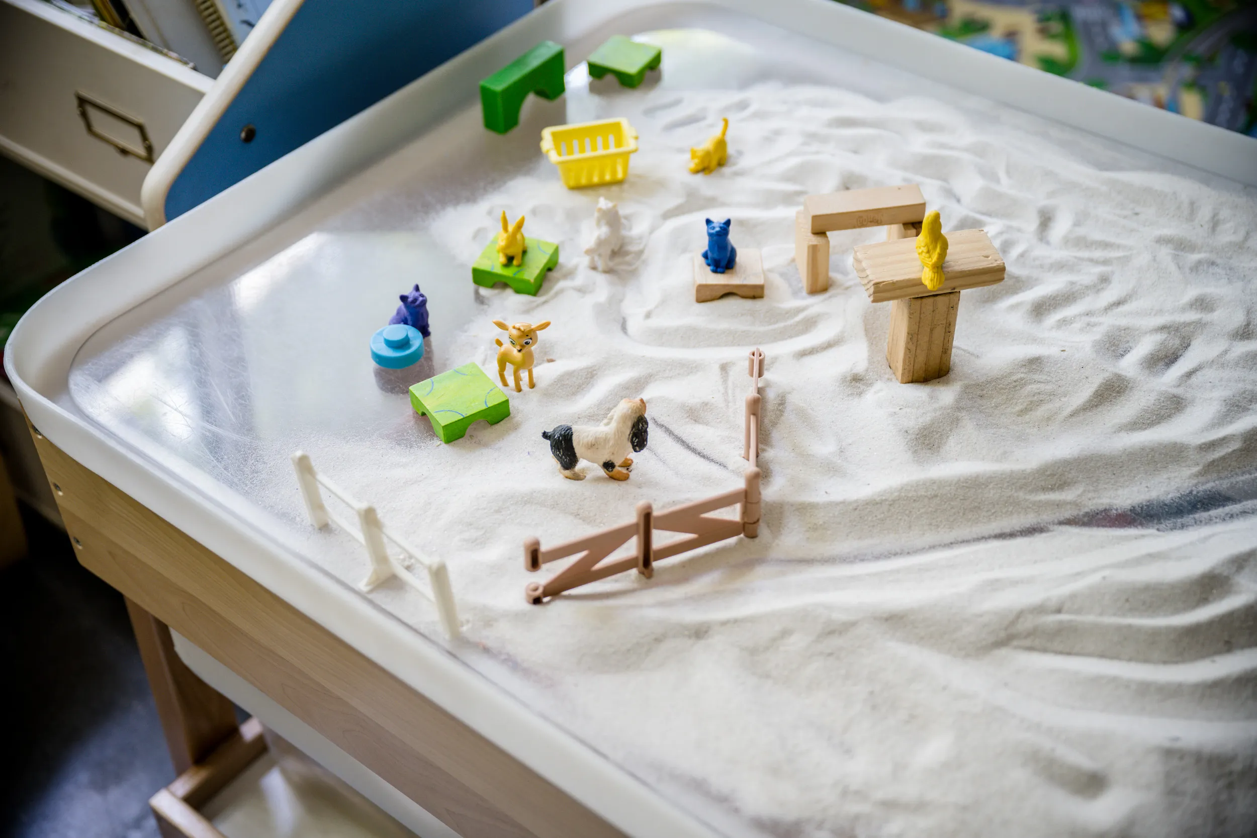 Une table sensorielle remplie de sable blanc accueille de petits animaux en plastique, des clôtures et des accessoires colorés, créant ainsi une scène de ferme miniature.