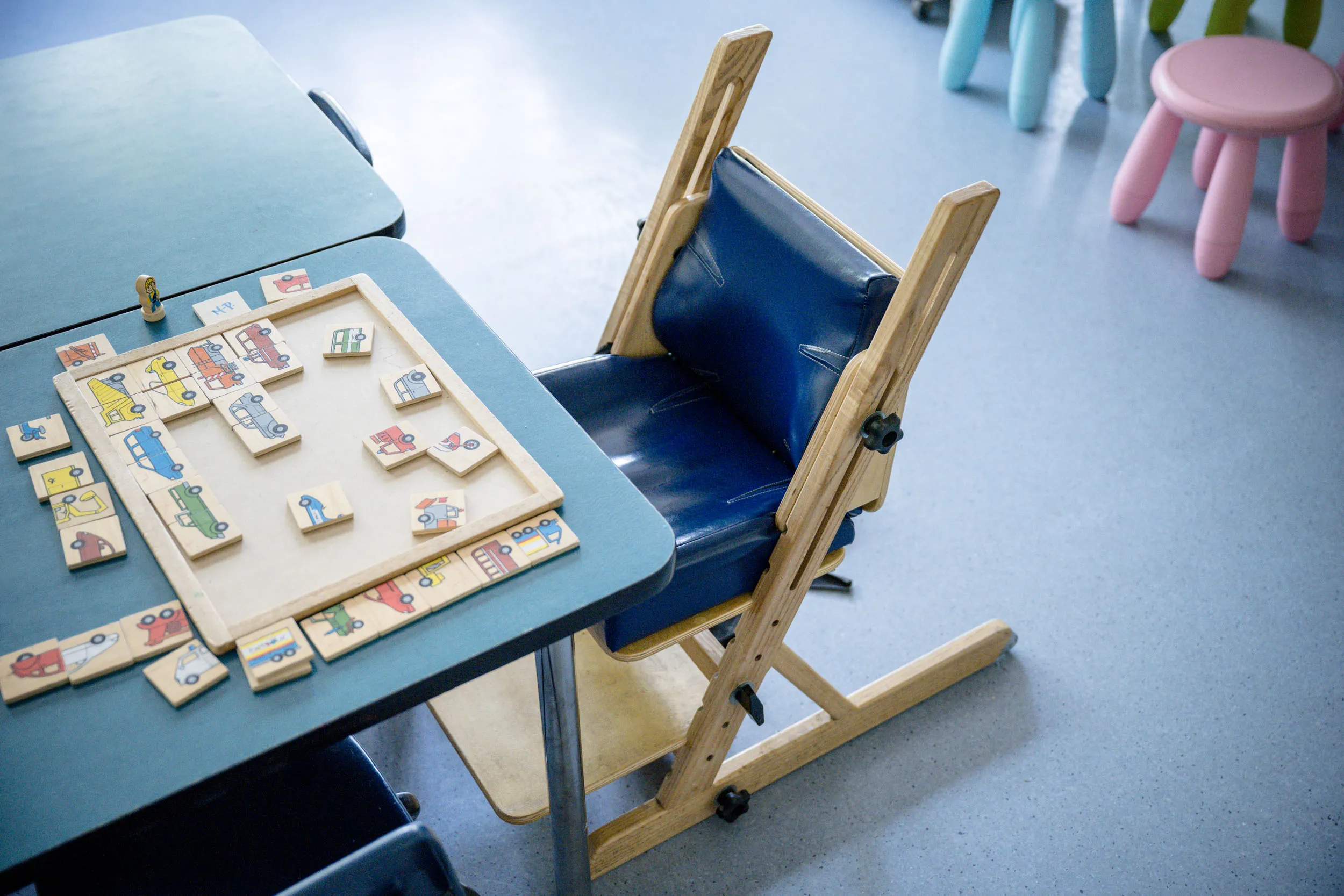 Une chaise adaptable bleue est posée près d'une table, à côté d'un puzzle composé de pièces en bois illustrées. À l'arrière-plan, sur un sol bleu clair, on aperçoit des meubles pour enfants aux couleurs vives.