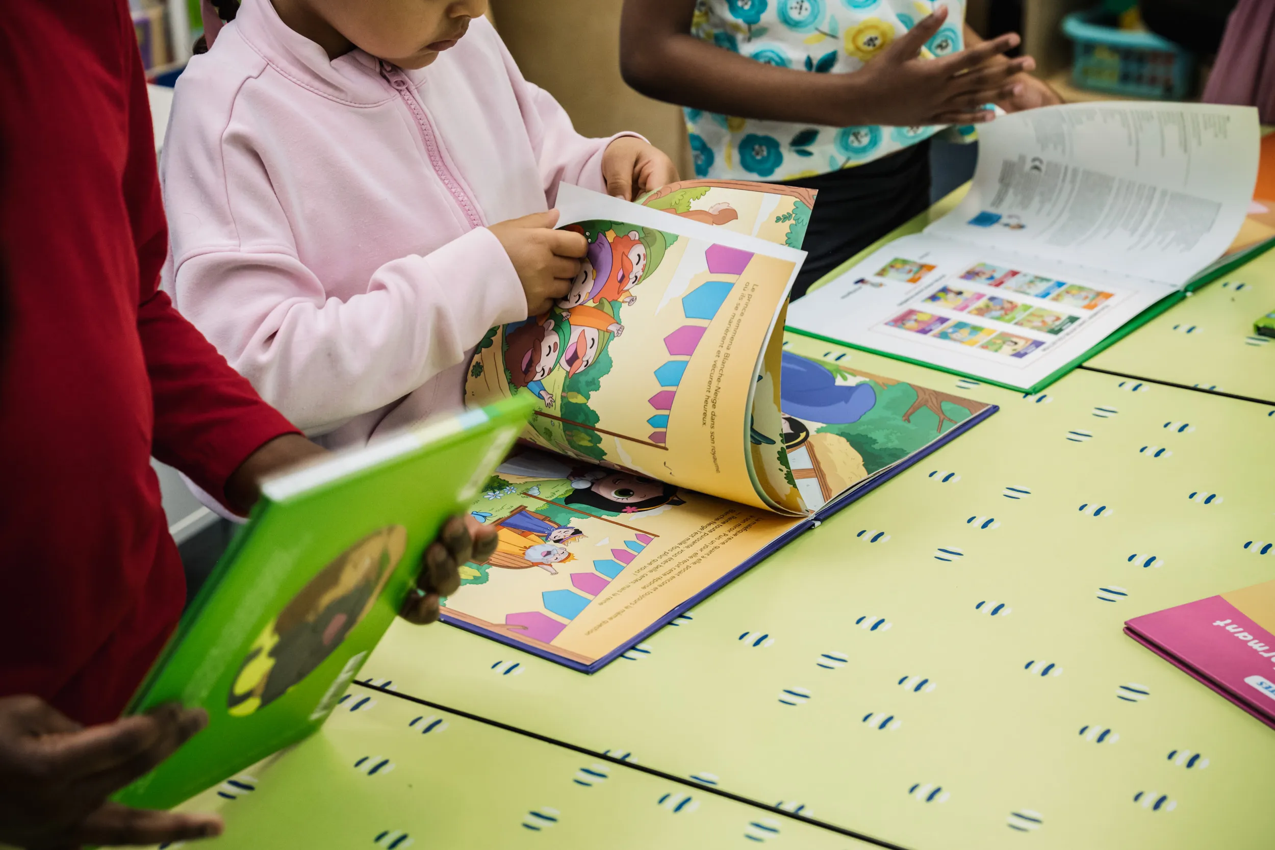 Trois enfants sont debout près d'une table, en train de lire et de feuilleter des livres d'images colorés. On ne voit que leurs mains et leur torse, et la table présente un plateau jaune clair à motifs.