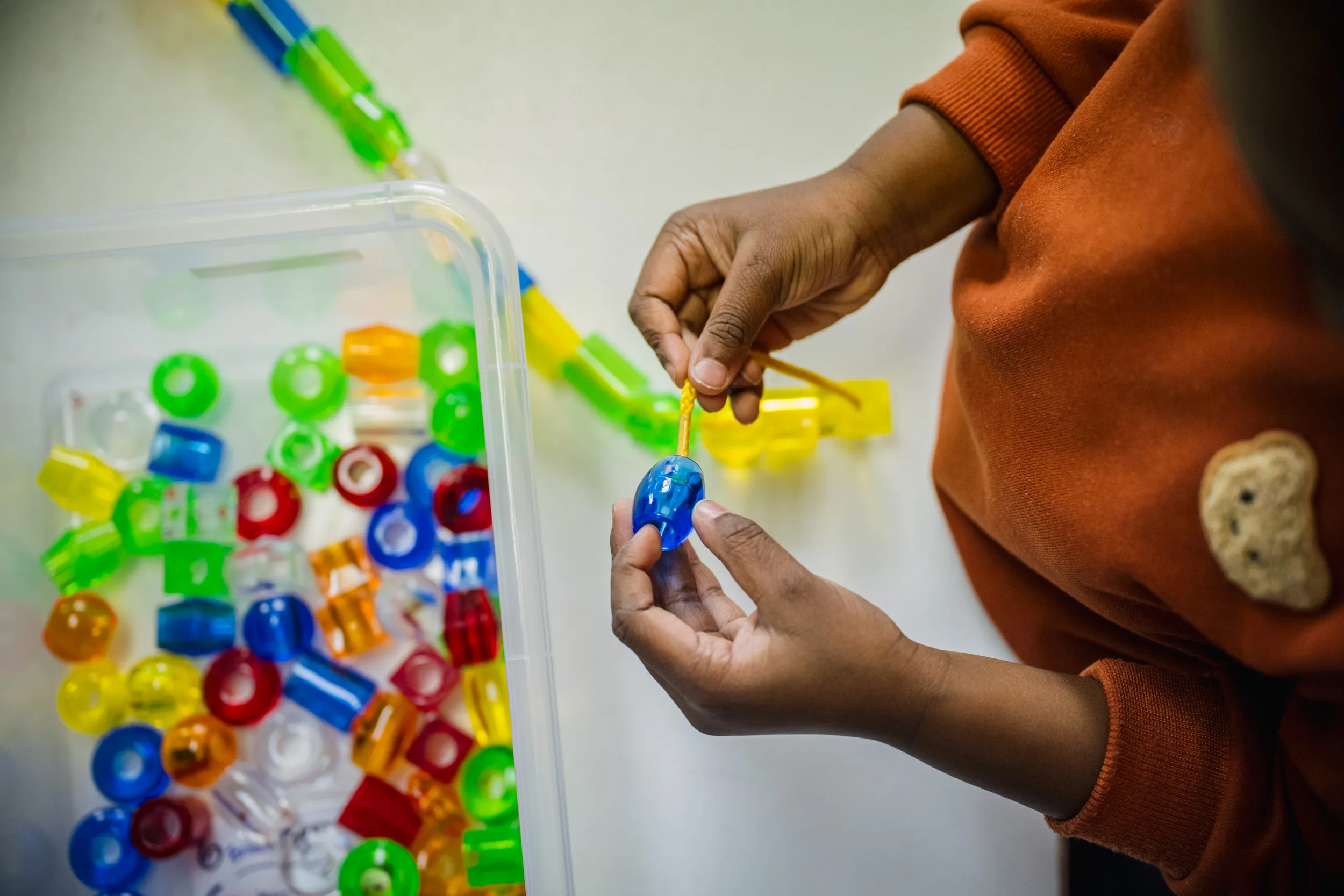 Un enfant vêtu d'un pull orange dispose des formes en plastique colorées et translucides sur une surface blanche, tenant une pièce bleue dans ses mains près d'un récipient transparent rempli de pièces similaires.