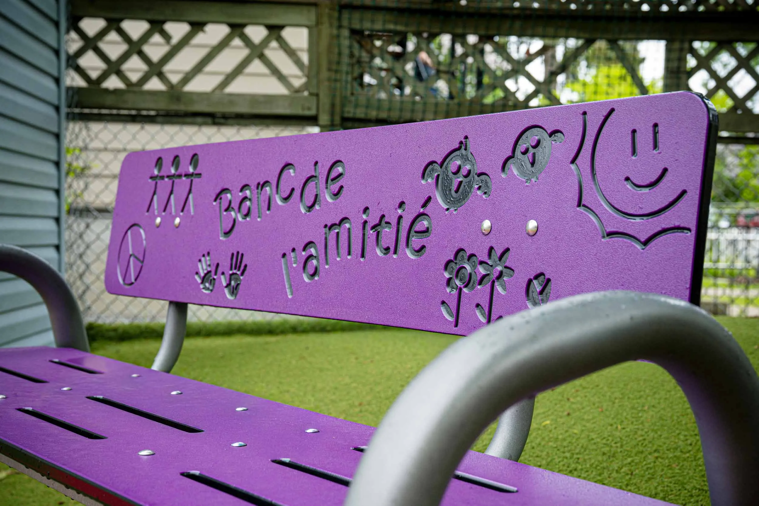 Un banc violet sur lequel sont gravés « BanC de lamitié » ainsi que des dessins joyeux représentant des fleurs, des smileys, des empreintes de mains et des symboles de paix sur le dossier. Le banc se trouve sur du gazon artificiel vert, près d'une clôture.